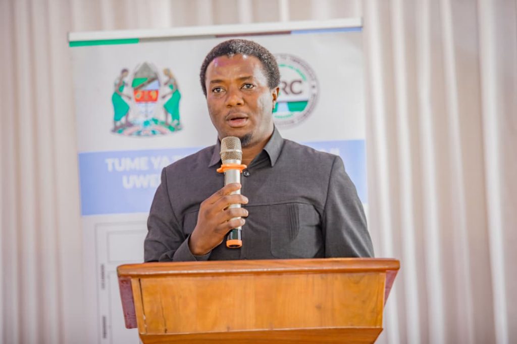 A man speaking at a podium with a microphone, wearing a black shirt, in front of a background featuring a banner with a coat of arms and text in a formal setting.